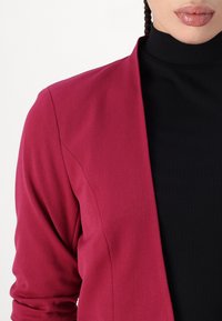 Přiléhavý kardigan s dlouhým rukávem v sytě magentové barvě, který má hladkou texturu a otevřený přední design, vrstvený přes černý žebrovaný top.