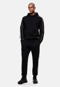 Sudadera negra con franjas laterales blancas, combinada con pantalones de chándal negros. Ambos hechos de una tela suave, con puños de canalé y un acento de logo.