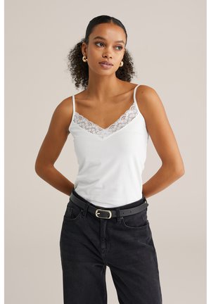 Biely top s krajkovým detailom pri výstrihu, s nastaviteľnými ramienkami, v kombinácii s tmavými čiernymi džínsami a čiernym opaskom.