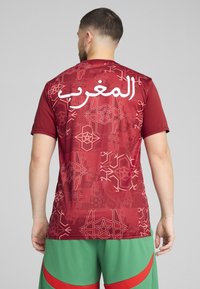 Cămașă sport roșie cu text alb în arabă și un model geometric, cu mâneci scurte și decolteu rotund.