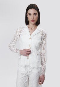 Chaqueta blanca de encaje con mangas largas, cierres de botones y un dobladillo en forma de escallola. Presenta intrincados patrones florales y una textura ligera.