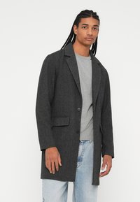 Grijs visgraat overjas met geknipte revers, sluiting met één knop en zijzakken, gedragen over een lichtgrijze trui en lichtdenim jeans.