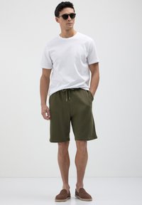 Homme portant des lunettes de soleil noires, un t-shirt blanc, un short à cordon olive et des chaussures à enfiler marron, se tenant avec une main dans sa poche.
