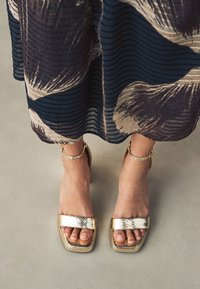 Aranyszínű, magas sarkú szandál strukturált kígyóbőr hatással, négyzet alakú orral és bokapánttal, egy könnyed, mintás ruhával párosítva.