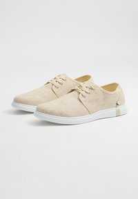 Zapatos de lona beige con suelas de goma blanca, diseño de cordones, punta redondeada, parte superior texturizada y pequeño detalle decorativo en el talón.