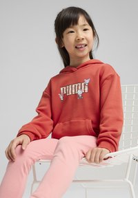 Koraldžasti dukserica s grafičkim printom na kojem su likovi i tekst "PUMA", uparen s svijetlopinkastim tajicama, sjedi na bijeloj žičanoj stolici.