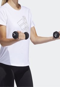 Hvid atletikshirt med sort Adidas-logo, sorte håndvægte holdt i begge hænder og sorte leggings. Tekstureret stof med korte ærmer.