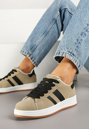 Baskets beiges avec des rayures noires et des lacets noirs épais portées avec un jean bleu clair sur un sol gris.