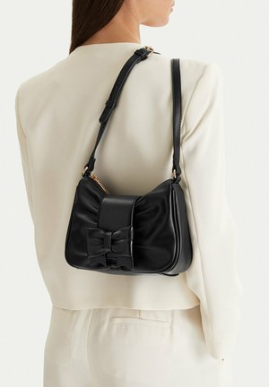 Mujer con un traje color marfil y un bolso de hombro de cuero negro con un gran lazo y detalles en dorado.