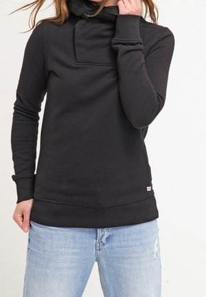 Czarna bluza z wysokim kołnierzem, z przednią kieszenią i prążkowanymi mankietami. Materiał wydaje się miękki i lekko teksturowany. Noszona z jasnoniebieskimi dżinsami.
