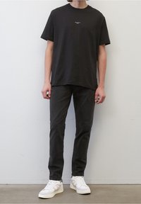 Sort bomulds T-shirt med rund hals og lille logo foran, parret med mørkegrå bukser og hvide sneakers. Minimalistisk design.