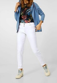 Blauwe zip-up hoodie, zwarte shirt met kleurrijk tropisch patroon, witte jeans en beige high-top sneakers met lichtblauwe veters.