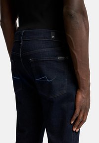 Jeans in denim blu scuro con un design aderente. Le caratteristiche includono cuciture a contrasto sulle tasche e un'etichetta brandizzata vicino alla vita sul retro.