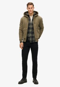 Sudadera con cremallera verde oliva, superpuesta sobre una camisa de cuadro, combinada con jeans negros y zapatos de cordones negros. Tejido suave con un corte casual.