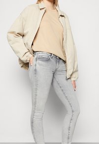 Chaqueta bomber beige, camiseta de tono claro y jeans skinny gris claro. La chaqueta cuenta con puños elásticos; los jeans son ajustados con un ligero desgaste.