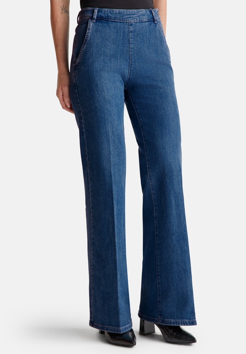 Femme portant un jean bleu large taille haute avec des poches latérales et des bottes noires à talons pointus, se tenant sur un fond blanc.