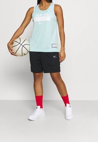 Mintgrön mesh-basketbollstopp med texten "FLY AS", tillsammans med svarta shorts. Innehåller även röda strumpor och vita sneakers, medan personen håller en basketboll.