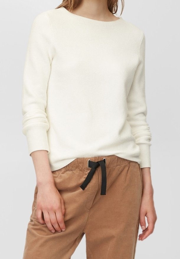 Femme portant un pull blanc uni à manches longues et un pantalon marron clair avec un cordon noir, debout sur un fond neutre.