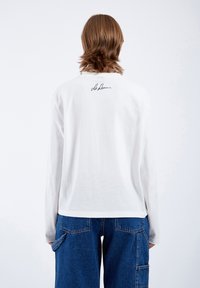 Long-sleeve white shirt with a round neckline, featuring a black signature detail on the back. Paired with loose blue denim pants.