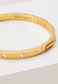 Goldfarbener Armreif mit glatter Oberfläche, der fünf gleichmäßig angeordnete klare Strasssteine und einen sicheren Verschluss bietet. Mit der Gravur "kate spade NEW YORK".