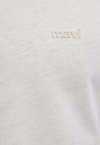 Weißer Strickstoff mit kleinem beigem "mavi"-Logo, das auf der oberen linken Seite gestickt ist.