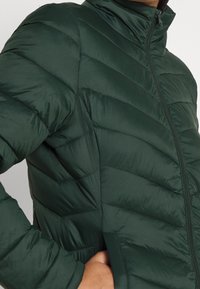 Tumma vihreä puffatakkia korkea kaulus, vaakasuora tikkauskuvio ja etuketju. Sileä, kiiltävä kangas ilman näkyviä logoja.