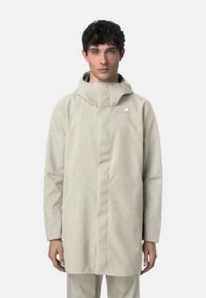 Giovane uomo che indossa un impermeabile beige chiaro con cappuccio, collo alto e logo sul petto, in piedi davanti a uno sfondo uniforme.