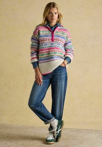 Strickpullover mit bunten geometrischen Mustern, geripptem Saum und Kragen mit Viertelreißverschluss; kombiniert mit lässigen blauen Jeans und grünen Sneakers.
