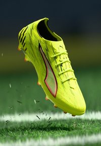Helles gelbes Fußballschuh mit strukturiertem Obermaterial, schwarzen Akzenten und roten Details. Verfügt über ein Schnürsystem und mehrere Stollen für besseren Halt.