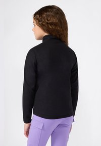 Bambino con capelli castani ricci fino alle spalle, indossa una giacca di pile nera e pantaloni cargo viola chiaro, rivolto a sinistra su uno sfondo neutro.