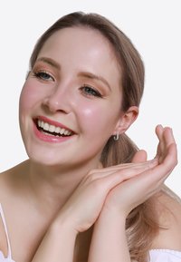Persona sorridente con capelli lisci, castano chiaro e un orecchino a cerchio argento, che mostra una pelle morbida e un trucco naturale. Mani uniti vicino al mento.