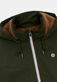 Grön jacka med hög krage, brun fleecefoder, en framzipper med röda och blå accenter, och dragsko vid halslinningen.