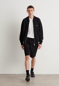 Schwarze Bomberjacke über einem weißen T-Shirt, kombiniert mit schwarzen gemusterten Shorts. Das Model trägt schwarze Socken und Sneaker. Einfaches Design, lässiger Look.