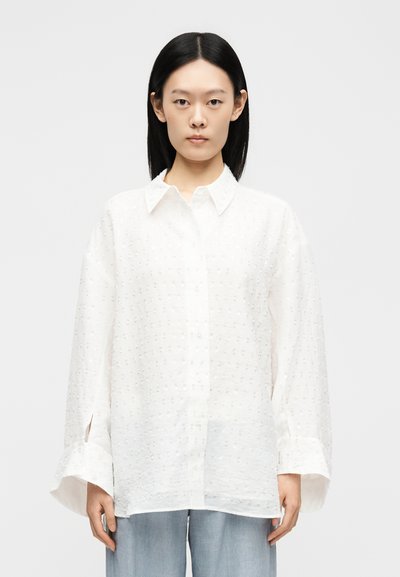 Femeie cu păr drept negru poartă o cămașă albă texturată, largă, cu nasturi și blugi albastru deschis, pe un fundal simplu.