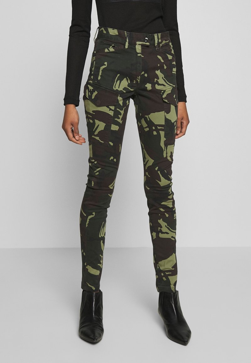 Pantalons skinny camouflage en tissu vert et marron avec une texture subtile. Présentent des poches avant et une fermeture éclair. Portés avec des bottines noires à talons pointus.