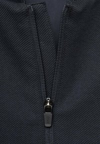 Prenda negra con cremallera, fabricada en un tejido texturizado, con un tirador de cremallera suave y costuras en contraste a lo largo de la línea de la cremallera.