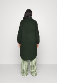 Oversized dark green coat with a back hem. Paired with houndstooth patterned green and white pants. Smooth texture and long sleeves.