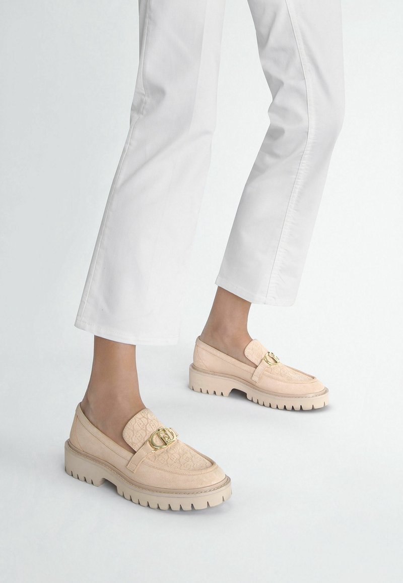 Pieds portant des mocassins beiges avec des accents de boucle dorée, associés à un pantalon blanc raccourci sur un fond clair uni.