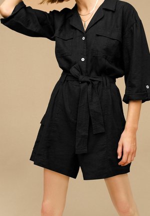 Femme portant une combinaison noire ceinturée avec des manches longues retroussées et deux poches poitrine sur un fond beige uni.