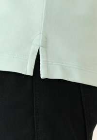 Top de algodón de color verde menta claro con un dobladillo curvado y aberturas laterales, combinado con una tela negra oscura por debajo. Costuras detalladas a lo largo de los bordes.