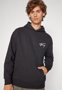 Felpa nera con cappuccio realizzata in morbido tessuto, dotata di una tasca frontale e polsini a costine. Include un logo bianco sul petto. Design semplice e casual.