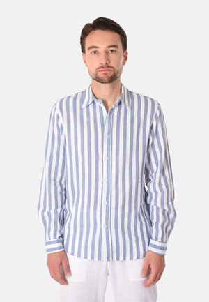 Uomo che indossa una camicia a maniche lunghe con righe verticali blu e bianche e pantaloni bianchi, in piedi davanti a uno sfondo chiaro e uniforme.