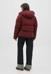 Burgundská zateplená bunda s kapucí, která má prošívaný design a volný střih, doplněná tmavými džíny s kontrastními manžetami.