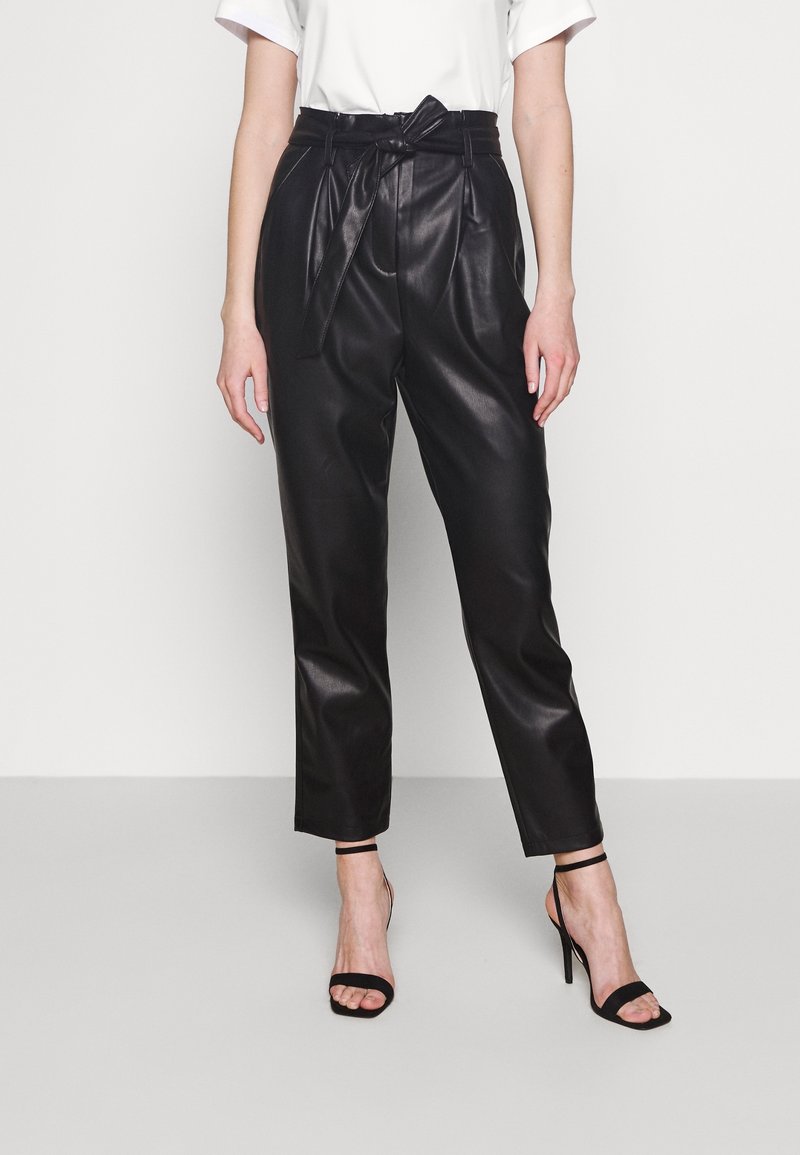 Pantalones de piel sintética negros con pliegues, cintura con cinturón y pierna ajustada. Combinados con tacones negros con tiras. Top blanco sencillo.