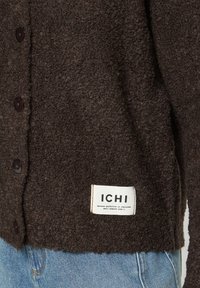 Gros plan d'une veste boutonnée texturée brun foncé avec une étiquette en tissu blanc ICHI et un jean en denim bleu en bas.