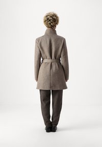 Manteau marron en mélange de laine avec une taille ceinturée, un col haut et une coupe ajustée, porté avec un pantalon gris foncé. Texture lisse et motif subtil visibles.