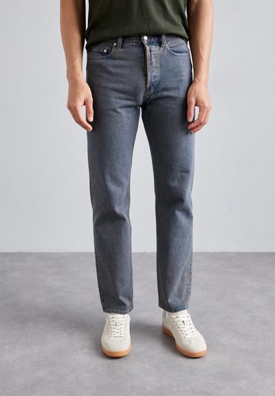 Blaue Jeans mit geradem Schnitt, ausgestattet mit fünf Taschen und Metallknopf-Beschlägen, kombiniert mit weißen Sneakern und einem grünen Oberteil.