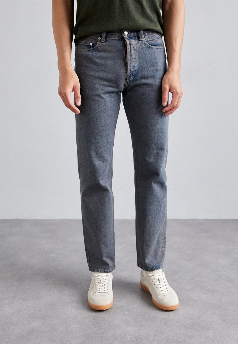 Vaqueros de mezclilla azul con corte recto, que cuentan con cinco bolsillos y herrajes de botón metálico, combinados con zapatillas blancas y una blusa verde.