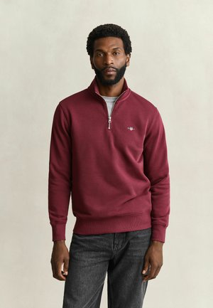 Mann mit kurzen lockigen Haaren und Bart, trägt einen bordeauxfarbenen Pullover mit Viertel-Reißverschluss und dunkelgraue Jeans, steht vor einem schlichten hellen Hintergrund.
