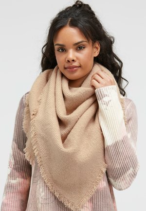Beige triangular scarf with fringed edges, made of textured fabric. Worn draped around the neck, paired with a multicolored patterned sweater.